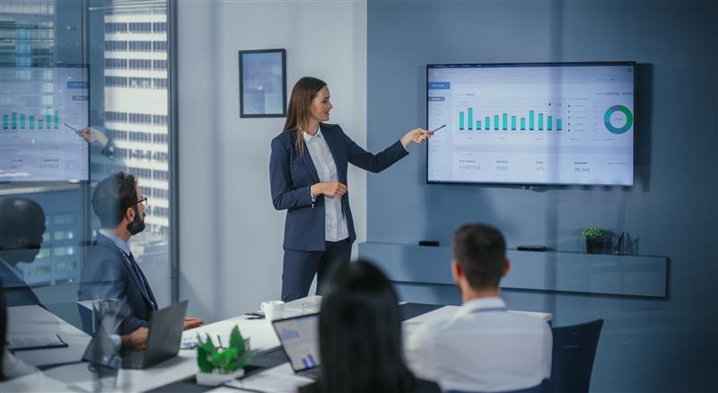 Businesswoman in a conference room presenting private equity financial charts on a wall-mounted screen to four seated colleagues, with bar graphs and a green pie chart visible on the display.